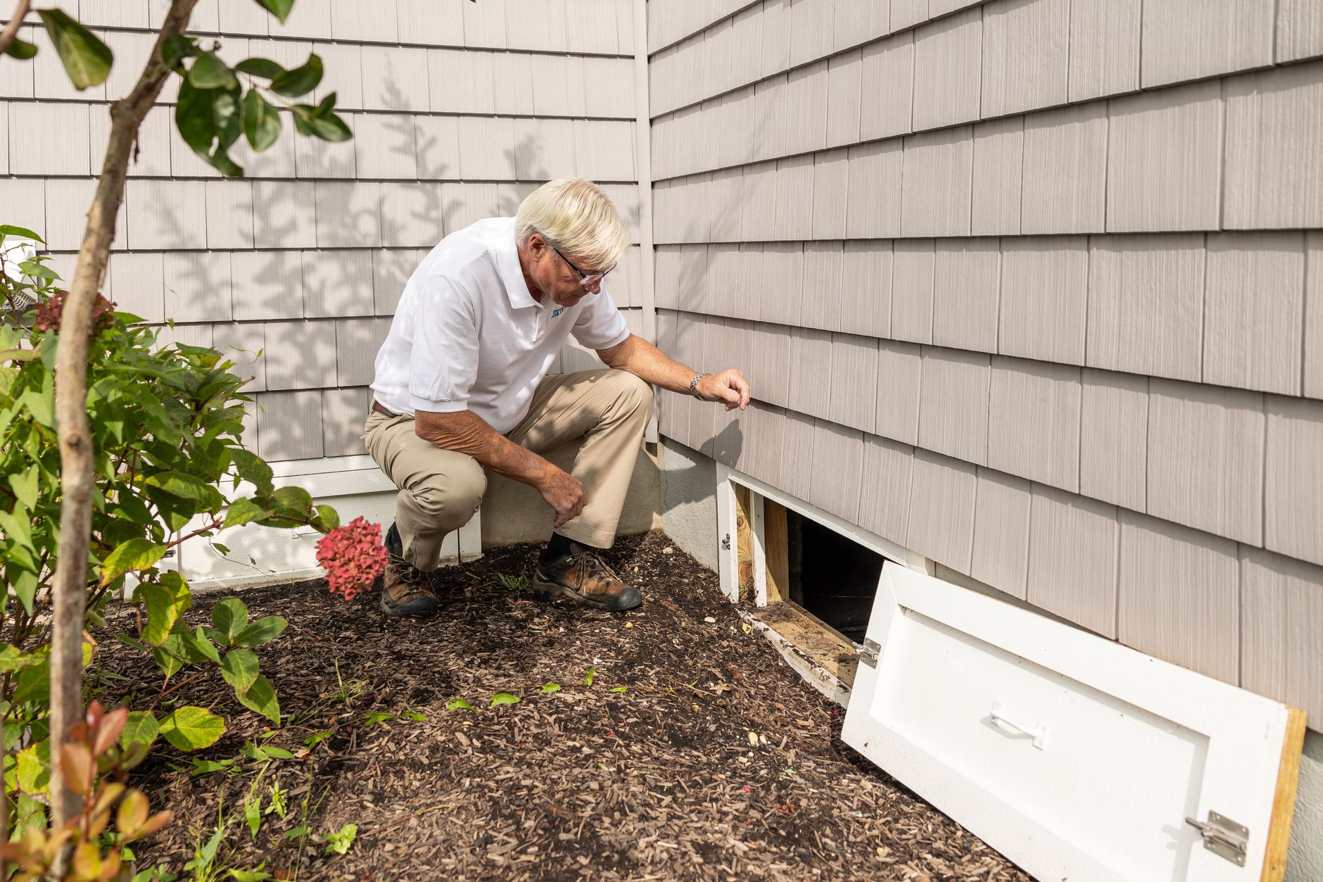 Senior adult inspecting a crawl space entrance during a home inspection on a sunny day.