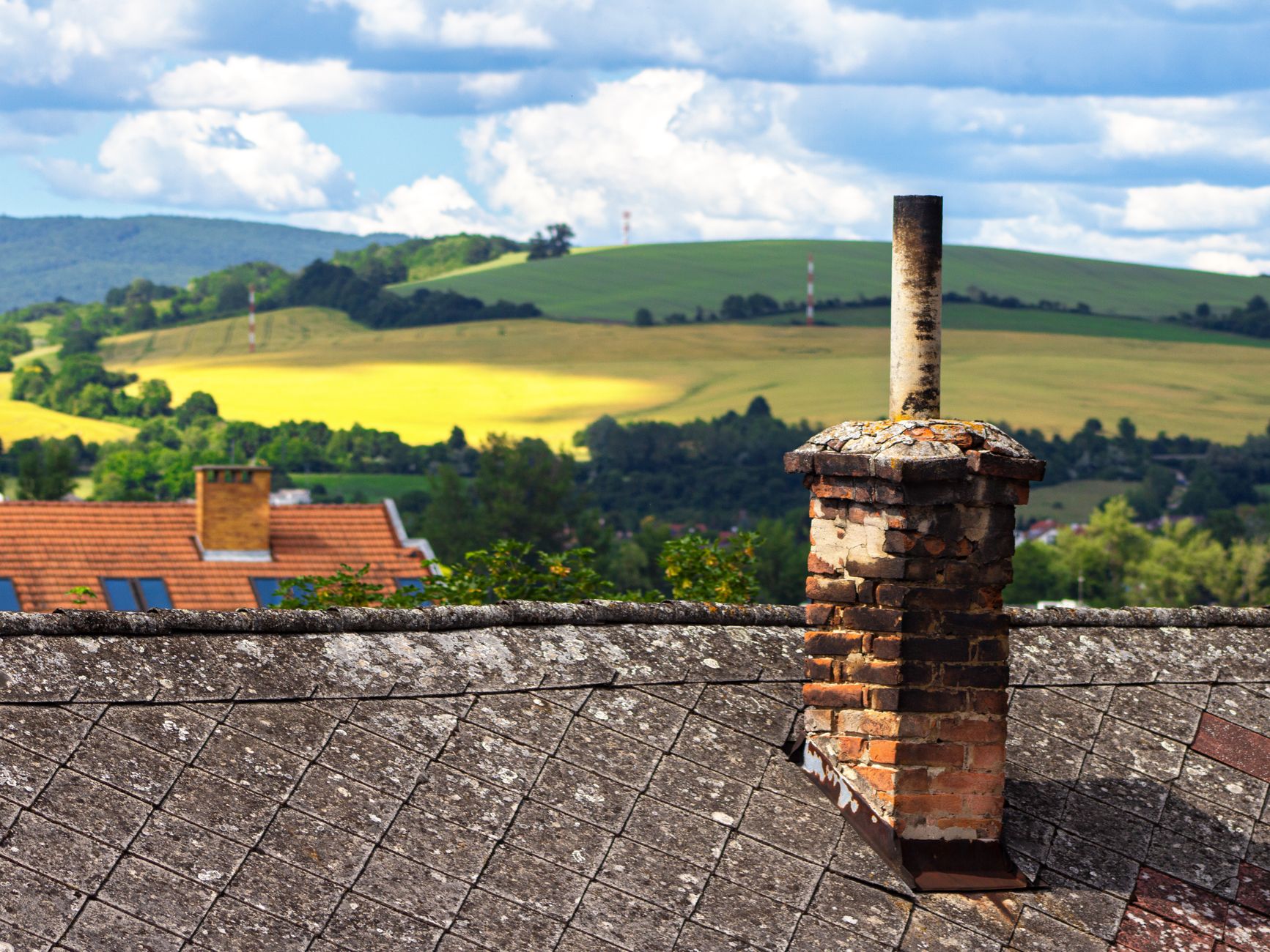 A rustic brick chimney on a weathered rooftop with rolling hills and fields in the background under a blue sky.